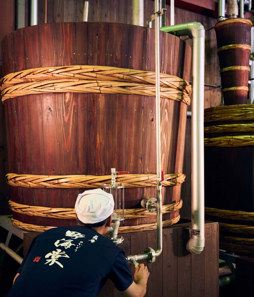 蔵人が木樽蒸留器の前で、蒸留している焼酎のアルコールチェックをしている様子